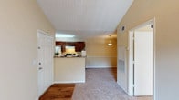This one-bedroom, one-bathroom unit at The Regency offers a thoughtfully arranged single-level layout with several notable architectural details. The living area immediately stands out with its vaulted ceilings, which lend a sense of openness and volume to the space. Warm beige walls and plush carpeting run throughout the main living and bedroom areas, while wood-style flooring accents the entryway and wet areas, creating a clean contrast.

The kitchen features a pass-through peninsula that connects it to the living space, offering a semi-open feel. Dark espresso-toned cabinetry lines both walls of the galley-style kitchen, complemented by granite-look countertops, a white appliance suite including a refrigerator and coil-top range, and a window that allows natural light in. A pendant light fixture above the peninsula adds a decorative touch to the dining area beyond.

The bedroom is generously proportioned, with a ceiling fan, a window overlooking greenery, and soft carpeting underfoot. The bathroom includes a full tub-shower combination with a curtained surround, dark wood cabinetry for storage, wood-style flooring, and clean white fixtures. Overall, the unit presents a cohesive, comfortable living environment with practical features throughout.
