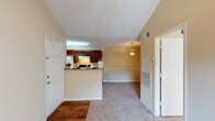 This one-bedroom, one-bathroom unit at The Regency offers a thoughtfully arranged single-level layout with several notable architectural details. The living area immediately stands out with its vaulted ceilings, which lend a sense of openness and volume to the space. Warm beige walls and plush carpeting run throughout the main living and bedroom areas, while wood-style flooring accents the entryway and wet areas, creating a clean contrast.

The kitchen features a pass-through peninsula that connects it to the living space, offering a semi-open feel. Dark espresso-toned cabinetry lines both walls of the galley-style kitchen, complemented by granite-look countertops, a white appliance suite including a refrigerator and coil-top range, and a window that allows natural light in. A pendant light fixture above the peninsula adds a decorative touch to the dining area beyond.

The bedroom is generously proportioned, with a ceiling fan, a window overlooking greenery, and soft carpeting underfoot. The bathroom includes a full tub-shower combination with a curtained surround, dark wood cabinetry for storage, wood-style flooring, and clean white fixtures. Overall, the unit presents a cohesive, comfortable living environment with practical features throughout.