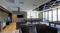 This stylish, single-floor space is designed as a sophisticated open-plan gathering area that seamlessly integrates a kitchen, lounge, and dining zone into one cohesive environment. The kitchen is anchored by a dark, substantial island with a built-in gas cooktop and a sleek industrial-style overhead range hood. A wall-mounted flat-screen television is positioned above the cooking area for added functionality and entertainment. Light hardwood flooring runs throughout the space, providing a warm contrast to the dark cabinetry and furnishings. The lounge area features a generously sized, textured sofa with decorative pillows, centered around a long, dark coffee table that adds to the room's modern aesthetic. A dramatic dining section accommodates a large group at an expansive table surrounded by upholstered chairs, lit by a striking chandelier-style pendant light. Floor-to-ceiling windows flood the dining area with natural light and frame sweeping urban views of the surrounding cityscape. An exposed geometric ceiling structure over the dining zone adds an industrial-chic architectural detail that elevates the overall design. The cohesive use of dark tones, recessed lighting, and contemporary furnishings throughout creates a refined yet functional ambiance ideal for entertaining or communal gatherings.