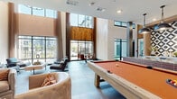 Located at 1320 Stanley St NE in Sugar Hill, Georgia, this contemporary two-story amenity lounge offers a well-appointed social and recreational space across two generously sized floors. The ground floor immediately impresses with soaring double-height ceilings and an expansive wall of floor-to-ceiling windows that flood the interior with natural light and provide open views of the surrounding community. A full-size pool table anchors the central game area, complemented by a stylish wet bar featuring a bold geometric-patterned tile accent wall, deep navy cabinetry, a granite-topped island with bar seating, and pendant lighting. A dedicated lounge zone showcases a linear fireplace framed in warm wood paneling, a wall-mounted TV, and tufted swivel chairs, creating an inviting gathering area. A tile-clad reception counter and large-format porcelain tile flooring reinforce the modern aesthetic throughout the lower level. The upper floor, accessible via an open staircase, continues the sophisticated design with a secondary lounge featuring a wet bar equipped with a wine cooler and beverage refrigerator, a wall-mounted TV, and additional seating areas. An open-railed mezzanine overlooks the double-height main floor below, connecting both levels visually and enhancing the sense of openness throughout the space.