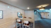 This thoughtfully designed single-floor space blends modern aesthetics with warm, natural materials to create a welcoming and functional environment. The standout feature of the space is a stylish high-top dining area framed by a striking light wood shiplap accent wall, which adds texture and warmth to the interior. The table is paired with contemporary white metal and brown leather bar-height chairs, providing a casual yet polished setting. Recessed lighting throughout the space ensures consistent, even illumination across each area.

A wide, well-lit corridor connects the various zones of the space, finished with dark flooring that contrasts cleanly against the light-toned walls. Modern flush-mount ceiling fixtures line the hallway, contributing to a clean and functional aesthetic. A modern staircase with a sleek metal railing is visible, adding an architectural element that enhances the overall design.

Adjacent to the corridor, a bright and open lounge or common area is visible through large glass-paneled windows and doors, allowing natural light to flow through and maintain a sense of openness and connectivity between spaces. The lounge features hardwood flooring and is furnished with comfortable seating and dining furniture, making it ideal for both collaborative gatherings and relaxed use. Overall, the space presents a cohesive and contemporary design with a welcoming ambiance.