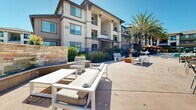 Located at 800 Reliant Way in American Canyon, California, this outdoor amenity space is designed to deliver a resort-style experience for its residents. The centerpiece is a generously sized swimming pool with crystal-clear water, framed by towering palm trees that lend a California resort ambiance. Adjacent to the pool, a bubbling spa provides a dedicated relaxation zone, complete with surrounding lounge chairs for sunbathing under the open sky.

The expansive pool deck is thoughtfully furnished with a variety of seating arrangements, including cushioned sectional sofas, lounge chairs, dining tables with shaded umbrellas, and white outdoor benches—all positioned to encourage both social gatherings and quiet relaxation. A pergola structure anchors one section of the courtyard, offering a shaded retreat.

The surrounding multi-story residential buildings feature stucco and wood panel exteriors accented by stone veneer detailing, large windows, and private balconies that overlook the amenity area. A gated entry with iron fencing ensures security and privacy. Mature landscaping with ornamental grasses, shrubs, and flowering plants adds color and greenery throughout, completing the inviting, well-maintained outdoor environment.