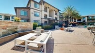 Located at 800 Reliant Way in American Canyon, California, this outdoor amenity space is designed to deliver a resort-style experience for its residents. The centerpiece is a generously sized swimming pool with crystal-clear water, framed by towering palm trees that lend a California resort ambiance. Adjacent to the pool, a bubbling spa provides a dedicated relaxation zone, complete with surrounding lounge chairs for sunbathing under the open sky.

The expansive pool deck is thoughtfully furnished with a variety of seating arrangements, including cushioned sectional sofas, lounge chairs, dining tables with shaded umbrellas, and white outdoor benches—all positioned to encourage both social gatherings and quiet relaxation. A pergola structure anchors one section of the courtyard, offering a shaded retreat.

The surrounding multi-story residential buildings feature stucco and wood panel exteriors accented by stone veneer detailing, large windows, and private balconies that overlook the amenity area. A gated entry with iron fencing ensures security and privacy. Mature landscaping with ornamental grasses, shrubs, and flowering plants adds color and greenery throughout, completing the inviting, well-maintained outdoor environment.