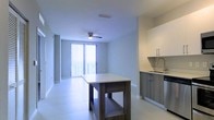 This single-floor space showcases a clean, modern design with an open-concept layout that seamlessly connects the kitchen and living area. The kitchen is outfitted with contemporary two-tone cabinetry featuring sleek hardware, a stainless steel sink, and a dishwasher, complemented by light-toned countertops. A freestanding wooden island with a lower storage shelf anchors the space, providing both additional prep space and a natural visual divider between the kitchen and the main living area. Large-format porcelain tile flooring runs throughout, lending a bright and cohesive feel to the interior. The living area is enhanced by a ceiling fan with integrated lighting, contributing to both comfort and functionality. Floor-to-ceiling vertical blinds frame a set of sliding glass doors that open to a private balcony, allowing for abundant natural light to fill the space and offering outdoor access. The neutral color palette — defined by soft gray walls, white ceilings, and white trim — creates a versatile and airy ambiance. The overall design reflects a contemporary aesthetic with practical, well-considered finishes suitable for modern living.