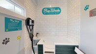 Pup Wash is a purpose-built pet grooming facility designed exclusively for the care of dogs and cats. The space is thoughtfully laid out on a single floor, combining functional amenities with a fun, welcoming atmosphere. The focal point of the room is a raised, walk-in pet bathing tub framed in a deep teal wood-panel surround, making it easy for pets to enter and exit comfortably. Floor-to-ceiling white subway tile lines the bathing area, creating a clean, hygienic environment that is both practical and visually crisp. A wall-mounted professional-grade pet dryer is positioned adjacent to the tub, providing a seamless grooming workflow. The walls throughout the space are decorated with colorful paw print decals in teal, yellow, and gray, accompanied by helpful grooming tips and instructional signage, adding an informative and cheerful character to the room. Protective aprons are conveniently hung near the grooming station for use during bath time. A glass door leads to an outdoor area, allowing natural light to filter into the space. Additional amenities include a step-on waste bin and safety equipment such as a fire extinguisher near the entrance. The durable flooring and easy-clean surfaces throughout reflect the practical design priorities of this well-equipped pet care station.