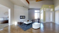 This contemporary single-floor space makes a striking impression with its soaring double-height ceilings and open, airy layout. The living area serves as the centerpiece, anchored by a sleek, horizontal linear fireplace set into a large, floor-to-ceiling white tile feature wall that adds a clean, minimalist aesthetic. Warm wood ceiling beams with track lighting provide a tasteful contrast to the otherwise modern palette of soft grays and light greens. Hardwood flooring runs throughout, lending warmth and cohesion to the space. Generous windows allow abundant natural light to pour in, brightening the open floor plan and highlighting the carefully considered design details. The living area comfortably accommodates a mix of modern furnishings, including chrome-frame chairs, a low-profile white coffee table, and a round white ottoman atop a plush blue area rug — all contributing to a clean, sophisticated atmosphere. A decorative potted plant adds a natural, organic touch to the interior. The overall ambiance is one of refined minimalism, where thoughtful architectural choices and an open layout come together to create a bright, welcoming, and stylish environment.