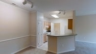 This single-floor space showcases an open layout that seamlessly connects the living area to the kitchen, creating a cohesive and functional environment. The living area features neutral-toned carpet, a two-tone wall treatment with a chair rail, and crown molding that adds a subtle architectural detail. Track lighting is mounted on the ceiling, providing flexible and adjustable illumination throughout the space.

The kitchen is defined by a curved peninsula with a dark countertop, offering additional workspace and the potential for casual seating. Light wood cabinetry lines the kitchen walls, complemented by a white electric range and an under-cabinet range hood. A skylight-style flush ceiling fixture brightens the kitchen area with additional overhead light. The kitchen also benefits from track lighting, ensuring the workspace remains well-lit.

Multiple paneled white doors lead to additional rooms or storage areas, contributing to the overall functionality of the layout. The neutral color palette of warm beige tones throughout the walls, flooring, and trim gives the space a clean, understated aesthetic that is both practical and welcoming.