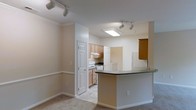 This single-floor space showcases an open layout that seamlessly connects the living area to the kitchen, creating a cohesive and functional environment. The living area features neutral-toned carpet, a two-tone wall treatment with a chair rail, and crown molding that adds a subtle architectural detail. Track lighting is mounted on the ceiling, providing flexible and adjustable illumination throughout the space.

The kitchen is defined by a curved peninsula with a dark countertop, offering additional workspace and the potential for casual seating. Light wood cabinetry lines the kitchen walls, complemented by a white electric range and an under-cabinet range hood. A skylight-style flush ceiling fixture brightens the kitchen area with additional overhead light. The kitchen also benefits from track lighting, ensuring the workspace remains well-lit.

Multiple paneled white doors lead to additional rooms or storage areas, contributing to the overall functionality of the layout. The neutral color palette of warm beige tones throughout the walls, flooring, and trim gives the space a clean, understated aesthetic that is both practical and welcoming.