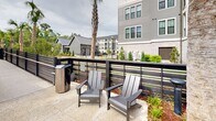 Located at 350 Henslow Dr in Charleston, SC, this outdoor amenity space offers a resort-style pool experience designed for relaxation and recreation. The centerpiece is a generously sized, freeform swimming pool with crystal-clear turquoise water, featuring gradual depth transitions and wide entry steps for easy access. The surrounding pool deck is finished in light-colored concrete and is outfitted with an array of lounge chairs and chaise lounges, many positioned beneath blue market umbrellas to provide shade throughout the day. A dedicated seating area with Adirondack-style chairs is situated along a slatted horizontal fence, offering a quieter retreat with views of the surrounding landscaping. Mature trees frame portions of the deck, providing natural shade and a lush, park-like atmosphere. A charming pool house structure with board-and-batten siding, a metal roof, and a covered porch with white railings anchors one end of the space, offering additional amenities and a welcoming architectural focal point. The backdrop features a multi-story residential building clad in gray siding and brick, with palm trees and manicured plantings enhancing the coastal Lowcountry aesthetic. The overall design blends contemporary outdoor living with the natural beauty of the Charleston landscape.