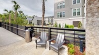 Located at 350 Henslow Dr in Charleston, SC, this outdoor amenity space offers a resort-style pool experience designed for relaxation and recreation. The centerpiece is a generously sized, freeform swimming pool with crystal-clear turquoise water, featuring gradual depth transitions and wide entry steps for easy access. The surrounding pool deck is finished in light-colored concrete and is outfitted with an array of lounge chairs and chaise lounges, many positioned beneath blue market umbrellas to provide shade throughout the day. A dedicated seating area with Adirondack-style chairs is situated along a slatted horizontal fence, offering a quieter retreat with views of the surrounding landscaping. Mature trees frame portions of the deck, providing natural shade and a lush, park-like atmosphere. A charming pool house structure with board-and-batten siding, a metal roof, and a covered porch with white railings anchors one end of the space, offering additional amenities and a welcoming architectural focal point. The backdrop features a multi-story residential building clad in gray siding and brick, with palm trees and manicured plantings enhancing the coastal Lowcountry aesthetic. The overall design blends contemporary outdoor living with the natural beauty of the Charleston landscape.