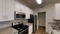 This single-floor residence offers a clean, contemporary layout designed for comfortable everyday living. The kitchen serves as a central highlight of the space, showcasing white shaker-style cabinetry that extends to the upper walls, providing generous storage throughout. Granite-style countertops add a polished, durable surface for meal preparation, while a full suite of stainless steel appliances — including a refrigerator, electric range, and over-the-range microwave — delivers a cohesive, modern aesthetic. A peninsula counter with a raised breakfast bar separates the kitchen from the adjacent living area, creating an open, connected feel ideal for entertaining or casual dining. The warm-toned hardwood-style flooring runs continuously through the kitchen and living areas, grounding the space with texture and visual warmth. Neutral gray walls complement the bright cabinetry and contribute to the light, airy atmosphere. Overhead flush-mount and fluorescent lighting ensures the kitchen and living areas remain well-lit. A hallway visible from the kitchen leads to additional rooms, suggesting a thoughtful and functional floor plan. Overall, the space presents a move-in-ready environment with modern finishes throughout.