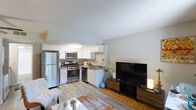 This single-floor residence offers a seamlessly connected open-concept layout that combines the kitchen and living area into one cohesive, functional space. The kitchen is well-appointed with a full suite of stainless steel appliances, including a refrigerator, gas range, built-in microwave, and dishwasher. Crisp white cabinetry lines the walls, providing ample storage, while the countertops and sink are positioned to maximize workflow efficiency. Light-toned laminate flooring runs throughout, adding warmth and visual continuity across the space. The living area flows naturally from the kitchen, offering a comfortable setting accented by a warm-toned area rug and a sleek media console. A textured ceiling adds subtle character overhead, while neutral-toned walls keep the atmosphere bright and versatile. The overall design balances modern practicality with a welcoming ambiance, making it a functional and inviting space suited for everyday living.