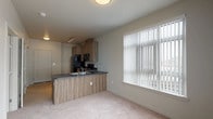 This single-floor unit presents a contemporary design with a practical, open-concept layout. The kitchen is a standout feature, outfitted with wood-grain cabinetry in a warm walnut-toned finish stacked to the ceiling for ample storage. Black Whirlpool appliances—including a refrigerator, electric range, over-the-range microwave, and dishwasher—are integrated throughout. Dark charcoal countertops provide a strong contrast against the cabinetry, and the kitchen flows seamlessly around an L-shaped peninsula with an undermount double sink. Light wood-style flooring runs through the kitchen and entry areas, grounding the space with a clean, modern aesthetic. Track lighting illuminates the kitchen zone.

The adjacent living area is carpeted and benefits from a large window fitted with vertical blinds, allowing for generous natural light. The bedroom is similarly carpeted, with its own sizeable window overlooking the exterior, and features a paneled interior door with built-in ventilation panels.

The bathroom is efficiently designed with a single vanity, dark countertop, an oval undermount sink, and a wide mirror accompanied by overhead lighting. The unit's overall palette is neutral—whites, greys, and warm wood tones—creating a cohesive and move-in-ready environment.
