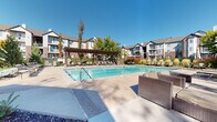 This thoughtfully designed outdoor amenity space serves as a central gathering hub for its residential community. The centerpiece is a generously sized swimming pool with crystal-clear water, surrounded by an expansive concrete deck furnished with lounge chairs and shaded by a handsome wooden pergola — ideal for relaxing on sunny days. Adjacent to the pool, a comfortable outdoor lounge area features wicker seating arranged around a striking circular fire pit crafted from natural river rock, providing a warm and inviting atmosphere for evening gatherings. The community grounds are attractively landscaped with ornamental grasses, mature trees, and manicured shrubs that add seasonal color and texture throughout the year. A dedicated hot tub enclosure, finished with distinctive river rock stonework on its exterior walls and large windows that flood the interior with natural light, offers a semi-private retreat. Inside, tile wainscoting and wall-mounted sconces complement the tiled hot tub, while views of the surrounding greenery and playground are visible through the windows. The architecture throughout features a blend of stone accents, wood detailing, and neutral stucco finishes, creating a cohesive and appealing aesthetic. The overall ambiance is relaxed, resort-inspired, and well-suited for both active recreation and quiet leisure.