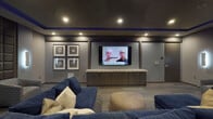 This single-floor media lounge is designed with a cinematic aesthetic, combining refined finishes and purposeful layout to create an immersive viewing and social environment. The room is anchored by a large wall-mounted flat-screen television positioned above a built-in cabinet with a granite or stone countertop, offering both display and storage functionality. Flanking the TV wall, a grid of framed artwork adds a polished, gallery-like touch to the space.

The seating arrangement centers on a generously sized navy blue sectional sofa adorned with neutral and gold accent pillows, complemented by a round upholstered ottoman at its center. Additional patterned swivel chairs are positioned near a dramatic floor-to-ceiling padded accent wall, which serves as a striking focal point and also provides acoustic benefits.

Vertical backlit sconces on either side of the accent wall contribute to the moody, layered lighting scheme, which is further enhanced by vivid blue LED cove lighting that traces the ceiling perimeter. Recessed lighting throughout ensures even illumination across the room. The carpeted flooring adds comfort underfoot while softening acoustics. Overall, the space exudes a dark, luxurious ambiance well-suited for entertainment and relaxation.