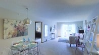This single-floor residence features an open-concept layout that seamlessly connects the dining and living areas, creating a spacious and cohesive flow throughout the main living space. The dining area is anchored by a sleek glass-top table with acrylic chairs and a contemporary spherical pendant light overhead, accompanied by colorful wall art that adds a vibrant focal point. Adjacent to the dining space, a large mirror enhances the sense of depth and light in the room. The living area is finished with plush carpeting and is furnished with a comfortable seating arrangement centered around an area rug. A wall-mounted television and a decorative console contribute to the functional yet stylish setup. Natural light fills the space through a large sliding glass door that opens onto a private balcony, offering views of the surrounding area. The textured ceiling adds a classic architectural detail throughout. A doorway visible from the main living area leads into a bedroom, which features its own window, continuing the bright and airy character of the home. Overall, the space presents a clean, well-lit interior with a practical and inviting layout.