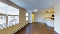 Located at 3 Chelsea in Cumberland Foreside, Maine, this single-floor residence offers a thoughtfully designed layout that balances openness with functionality. The main living area is characterized by rich, dark hardwood flooring that extends throughout, creating a warm and cohesive aesthetic. Neutral beige walls provide a clean, versatile backdrop, while multiple windows fitted with white blinds allow natural light to fill the space, offering views of the surrounding neighborhood. A baseboard heating system runs along the walls, ensuring year-round comfort. The open floor plan connects the living area seamlessly to the kitchen, which features white cabinetry and a light-toned countertop visible along the far edge of the space. A white entry door with a deadbolt provides secure access and is positioned at the rear of the main area, with a hallway leading to additional rooms. Flush-mount ceiling lighting provides consistent illumination throughout. The overall ambiance is clean, bright, and move-in ready, with a straightforward design that highlights practical finishes and a welcoming sense of space suitable for everyday living in the Cumberland Foreside community.