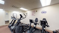 Located at 111 N Fourth St in downtown Columbus, Ohio, this single-floor fitness facility offers a practical and motivating environment for exercise. The gym is organized into distinct functional zones, making efficient use of the available floor space.

The strength training area is anchored by a Paramount SL multi-station cable machine, a robust piece of equipment supporting a wide range of resistance exercises. Complementing this is a rack of dumbbells in graduated weights, along with an adjustable bench, providing a thorough free-weight training setup. The flooring in this section features low-pile carpet, and vertical blinds cover a nearby window.

The cardio zone is well-appointed with multiple elliptical trainers and recumbent stationary bikes, each equipped with individual console displays. Motivational phrases are displayed prominently on the walls, including "The body achieves what the mind believes" and "Will it be easy? Nope. Worth it? Absolutely," contributing to an encouraging atmosphere. Recessed fluorescent panel lighting illuminates both areas via a drop-tile ceiling, maintaining consistent brightness throughout. Additional rooms in the facility, while minimally lit in available images, appear to include supplementary spaces with windowed walls fitted with horizontal blinds.
