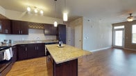 This single-story, 3-bedroom, 2-bathroom residence offers a well-designed open-concept layout that seamlessly connects the kitchen and living areas. The kitchen is a standout feature, showcasing rich dark-espresso cabinetry, granite countertops with warm gold and cream tones, and a large central island ideal for meal prep or casual dining. Pendant lighting above the island adds a refined touch, complementing the track lighting overhead. A built-in wine rack integrated into the upper cabinetry provides a distinctive and functional design detail. The kitchen is equipped with a double-basin sink, dishwasher, and ample storage throughout. Warm hardwood flooring flows continuously through the kitchen and into the adjoining living space, creating a cohesive and inviting ambiance. The open living area offers generous space with neutral wall tones that lend a clean, versatile backdrop. Ceiling vents throughout ensure efficient climate control. The overall design balances practicality with modern aesthetics, making this home well-suited for both everyday living and entertaining.