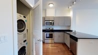 This single-floor space is designed with a clean, contemporary aesthetic that prioritizes both function and style. The kitchen serves as a central highlight, featuring stainless steel appliances including a range, built-in microwave, and dishwasher, all complemented by dark granite-style countertops and modern flat-front cabinetry in a warm grey finish. Track lighting and a flush-mount ceiling fixture provide ample illumination, while rich hardwood flooring adds warmth and continuity throughout. The kitchen layout is thoughtfully configured in an L-shape, maximizing counter and storage space without sacrificing flow. A stainless steel sink is positioned along one side, offering practicality alongside the polished design. One of the unit's standout features is the in-unit laundry, with a full-size stacked washer and dryer neatly concealed behind classic louvered doors — a convenient and space-efficient solution. The overall ambiance is modern and well-appointed, reflecting a residence that balances everyday practicality with a refined, urban sensibility.