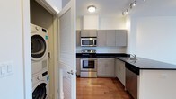 This single-floor space is designed with a clean, contemporary aesthetic that prioritizes both function and style. The kitchen serves as a central highlight, featuring stainless steel appliances including a range, built-in microwave, and dishwasher, all complemented by dark granite-style countertops and modern flat-front cabinetry in a warm grey finish. Track lighting and a flush-mount ceiling fixture provide ample illumination, while rich hardwood flooring adds warmth and continuity throughout. The kitchen layout is thoughtfully configured in an L-shape, maximizing counter and storage space without sacrificing flow. A stainless steel sink is positioned along one side, offering practicality alongside the polished design. One of the unit's standout features is the in-unit laundry, with a full-size stacked washer and dryer neatly concealed behind classic louvered doors — a convenient and space-efficient solution. The overall ambiance is modern and well-appointed, reflecting a residence that balances everyday practicality with a refined, urban sensibility.