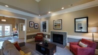 This single-floor space makes a strong first impression with its thoughtfully designed living area. The room is anchored by a striking fireplace framed in a mosaic tile surround, adding a refined focal point to the space. Soaring tray ceilings elevate the sense of height and grandeur, while crown molding and a classic chair rail running along the walls contribute a timeless, architectural elegance.

The walls are finished in warm neutral tones, complemented by crisp white trim that enhances the bright, welcoming ambiance. Recessed lighting is positioned throughout the ceiling, providing even illumination across the room. A built-in media niche with shelving offers both functional storage and a clean, integrated aesthetic.

The living area is furnished with plush seating in rich earth tones — including deep red and amber upholstered chairs — arranged around a substantial dark wood coffee table. Framed artwork adorns the walls, adding color and personality to the space. Natural light filters in through a glass-paneled door visible in the background, further brightening the interior. Soft area rugs and carefully chosen décor complete the look, making this a comfortable, stylish, and cohesive living environment.