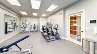 Located at 155 Windermere Ave in Ellington, Connecticut, this well-equipped fitness center offers a clean, modern environment designed to support a full range of workout routines. The single-floor space is bathed in natural light from multiple large skylights set into the ceiling, complemented by recessed panel lighting that ensures consistent brightness throughout the room. Neutral grey flooring and soft blue-grey walls create a calm, focused atmosphere.

The cardio zone features multiple treadmills and an elliptical trainer, all positioned near windows with slatted blinds that allow natural light to filter in. A recumbent bike and upright cycle provide additional low-impact options. The strength training area is outfitted with a comprehensive set of Matrix-brand selectorized machines, including cable and pulley systems, as well as a leg extension and chest press unit. A multi-tiered dumbbell rack holds a full range of free weights, accompanied by an adjustable Matrix workout bench.

Large wall-mounted mirrors span one side of the room, visually expanding the space and supporting proper form during exercise. Shiplap-style white wall paneling adds a refined, contemporary design touch. A glass-paned door leads to an adjoining restroom facility, and decorative artwork adorns the walls, rounding out this thoughtfully designed community fitness space.