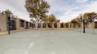 This outdoor recreational space is part of a multi-building residential complex characterized by its low-rise, stucco-clad structures featuring tiled rooflines and a neutral earth-toned exterior palette. The grounds are anchored by a full-size basketball court with clearly marked court lines and regulation backboards, enclosed within chain-link fencing alongside an adjacent tennis court. Concrete pathways wind throughout the property, connecting the various buildings and amenity areas. Mature deciduous trees are interspersed across the grounds, providing natural shade and visual character to the open landscape. The surrounding terrain consists largely of dry, patchy grass and gravel-covered areas, reflecting a dry climate environment. Exterior utility infrastructure, including PVC plumbing fixtures, is visible along the building walls. One image reveals a section of the building's exterior wall with exposed structural layers, indicating areas undergoing maintenance or repair. Pole-mounted lighting fixtures are positioned throughout the grounds to provide illumination across the common areas. The overall layout is open and functional, designed to accommodate outdoor activity and pedestrian circulation throughout the property.