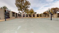 This outdoor recreational space is part of a multi-building residential complex characterized by its low-rise, stucco-clad structures featuring tiled rooflines and a neutral earth-toned exterior palette. The grounds are anchored by a full-size basketball court with clearly marked court lines and regulation backboards, enclosed within chain-link fencing alongside an adjacent tennis court. Concrete pathways wind throughout the property, connecting the various buildings and amenity areas. Mature deciduous trees are interspersed across the grounds, providing natural shade and visual character to the open landscape. The surrounding terrain consists largely of dry, patchy grass and gravel-covered areas, reflecting a dry climate environment. Exterior utility infrastructure, including PVC plumbing fixtures, is visible along the building walls. One image reveals a section of the building's exterior wall with exposed structural layers, indicating areas undergoing maintenance or repair. Pole-mounted lighting fixtures are positioned throughout the grounds to provide illumination across the common areas. The overall layout is open and functional, designed to accommodate outdoor activity and pedestrian circulation throughout the property.