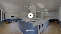 Located at 1500 Shadowridge Drive Southeast in Vista, CA, this single-floor community amenity facility combines a full-service laundry room with a dedicated fitness area in one efficiently designed space. The interior features a clean, modern aesthetic with a two-tone color scheme of crisp white upper walls and slate blue lower panels, complemented by light wood-style flooring throughout.

The laundry area is the centerpiece of the space, equipped with a generous row of front-load washing machines and dryers arranged along a central island and perimeter walls. A white laminate countertop surface spans the machine island, providing practical folding space. Overhead, bright flush-mount and recessed lighting ensures excellent visibility throughout the room.

A dedicated fitness alcove occupies one section of the facility, furnished with a recumbent exercise bike and an elliptical trainer, along with a wall-mounted flat-screen television — an ideal setup for staying active. Large windows line one wall of this area, flooding the space with natural light and offering pleasant views of an outdoor playground and surrounding greenery.

The facility's open floor plan promotes easy circulation, while the ample square footage accommodates multiple users simultaneously. Overall, the space delivers a functional, well-maintained environment designed to serve the everyday needs of a residential community.
