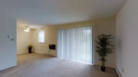 This single-floor space features a bright and open living area designed with simplicity and functionality in mind. The room is finished with wall-to-wall carpeting in a neutral tone, creating a comfortable and cohesive feel underfoot. A prominent wall of floor-to-ceiling sliding glass doors dominates one side of the room, dressed with full-length vertical blinds that allow for flexible light control while hinting at outdoor access beyond. Natural light also filters in through a secondary window on the adjacent wall, contributing to the overall brightness of the space. The walls are finished in a soft, warm beige tone that complements the light palette throughout. A baseboard heating unit runs along the lower portion of the wall beneath the window, providing a practical climate-control solution. The open layout offers generous room to arrange furnishings freely, making it a versatile living area. Decorative touches, such as a potted bamboo plant near the sliding doors, highlight the potential for a clean, welcoming aesthetic. Overall, the space presents a calm, understated atmosphere well-suited for comfortable everyday living.