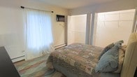 This single-floor bedroom offers a practical and comfortable layout designed for restful living. The room centers around a double bed dressed in a classic patchwork quilt, lending a warm, homey atmosphere to the space. Neutral, light-toned walls keep the room feeling bright and airy, complemented by natural light filtering through a curtained window dressed with sheer lace panels. A baseboard heater beneath the window ensures year-round comfort. A wall-mounted unit near the window provides additional functional convenience. The room features a notably generous closet area, with double bi-fold doors opening to reveal a spacious storage space — an asset for everyday organization. A second set of bi-fold doors suggests additional dedicated storage, offering ample room for clothing and belongings. The patterned area rug adds a subtle decorative touch to the floor, tying the room's cozy aesthetic together. Overall, the bedroom presents a straightforward, functional design with practical storage solutions, comfortable furnishings, and sufficient natural light, making it a welcoming and livable space.