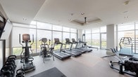 This fitness center is a thoughtfully designed, multi-zone facility that combines functionality with an elevated aesthetic. The space is defined by expansive floor-to-ceiling glass walls that flood the interior with abundant natural light and offer sweeping panoramic views of the surrounding cityscape and treetops, creating an energizing backdrop for workouts.

The cardio zone features a row of treadmills, elliptical trainers, and stationary bikes, including air resistance models, all positioned to face the dramatic window walls. A dedicated strength training area includes Cybex cable machines, a multi-function cable station, adjustable benches, and a dumbbell rack, complemented by a wall-mounted flat-screen television and large mirror panels that enhance the sense of depth. A separate section provides open floor space suitable for stretching and functional training, with rubber flooring for added safety and comfort.

Ceiling fans are integrated into tray ceiling details, contributing to air circulation and a refined architectural finish. Recessed lighting supplements natural light throughout. A corridor with textured blue-gray accent walls leads to private changing rooms or restroom facilities. An exterior glass door provides access to an outdoor area, further connecting the space to its elevated surroundings.