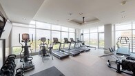 This fitness center is a thoughtfully designed, multi-zone facility that combines functionality with an elevated aesthetic. The space is defined by expansive floor-to-ceiling glass walls that flood the interior with abundant natural light and offer sweeping panoramic views of the surrounding cityscape and treetops, creating an energizing backdrop for workouts.

The cardio zone features a row of treadmills, elliptical trainers, and stationary bikes, including air resistance models, all positioned to face the dramatic window walls. A dedicated strength training area includes Cybex cable machines, a multi-function cable station, adjustable benches, and a dumbbell rack, complemented by a wall-mounted flat-screen television and large mirror panels that enhance the sense of depth. A separate section provides open floor space suitable for stretching and functional training, with rubber flooring for added safety and comfort.

Ceiling fans are integrated into tray ceiling details, contributing to air circulation and a refined architectural finish. Recessed lighting supplements natural light throughout. A corridor with textured blue-gray accent walls leads to private changing rooms or restroom facilities. An exterior glass door provides access to an outdoor area, further connecting the space to its elevated surroundings.