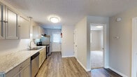 Located at 1600 Arlington Blvd in Greenville, NC, this single-floor unit offers a clean, updated interior designed for comfortable everyday living. The open living area is finished with durable wood-look plank flooring and features a ceiling fan with light fixture, providing both functionality and a welcoming ambiance. Textured ceilings and neutral wall tones carry throughout, creating a cohesive and bright aesthetic.

The kitchen is well-appointed with gray cabinetry, light granite-style countertops, a stainless steel sink, and stainless steel appliances including an electric range and refrigerator. A range hood is installed above the stove, and a door at the end of the kitchen provides direct exterior access, allowing natural light to filter in through its window.

Adjacent to the main living space, the bedroom features plush carpeting and a generously sized closet with double doors and a full-width hanging rod, offering ample storage. The bathroom is equipped with a full-size soaking tub and shower combo with a clean white surround.

A central hallway connects the key areas of the unit, with a louvered closet door providing additional storage. The overall layout is efficiently designed, making practical use of the available space while maintaining an open and airy feel throughout.