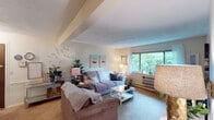 This single-floor residence offers a well-organized layout that balances comfort and functionality across its key living areas. The living room serves as the heart of the space, featuring a large window that frames lush greenery outside, filling the room with natural light. Soft carpeting, a textured ceiling, and an exposed beam detail contribute to its cozy, understated character. The room provides ample space for comfortable seating arrangements centered around a coffee table.

The kitchen is outfitted with dark espresso-toned cabinetry paired with light countertops, creating a classic contrast. It includes a double-basin sink, a freestanding range, and a window with a decorative mirror accent above the sink area. A pendant light adds a warm focal point, and additional shelving units provide practical storage and workspace nearby.

The bedroom is a tranquil retreat with plush carpeting, a wide window dressed in sheer curtains, and access to a walk-in closet with built-in shelving and drawer storage. The textured ceiling continues throughout, maintaining a consistent aesthetic.

The bathroom features a tub-shower combination with a curtain, a vanity with a marble-patterned countertop, a well-lit mirror, and over-toilet shelving for added storage. Overall, the space presents a warm, livable atmosphere with thoughtful details throughout.