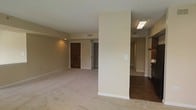 This single-floor residence offers a clean, open layout designed for comfortable everyday living. The main living area is finished with plush wall-to-wall carpeting in a warm neutral tone, complemented by smooth beige walls that create a bright and welcoming atmosphere. A structural column subtly divides the space, adding architectural character while maintaining an open feel throughout.

Adjacent to the living area, the transition to the kitchen zone is marked by rich hardwood-style flooring, providing a natural contrast to the carpeted spaces. The kitchen area is partially visible, hinting at cabinetry and functional prep space. Recessed and flush-mount lighting provides consistent illumination throughout the interior.

A wide entryway corridor leads to what appears to be additional rooms or closet space, suggesting a well-organized layout with thoughtful separation between living and private areas. The solid wood-paneled entry door adds a classic, grounded aesthetic to the space.

Throughout the residence, visible HVAC vents and clean baseboard trim reflect attention to practical detail. The overall ambiance is understated and versatile, offering a neutral canvas well-suited for a variety of interior styles and furnishing preferences.