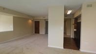 This single-floor residence offers a clean, open layout designed for comfortable everyday living. The main living area is finished with plush wall-to-wall carpeting in a warm neutral tone, complemented by smooth beige walls that create a bright and welcoming atmosphere. A structural column subtly divides the space, adding architectural character while maintaining an open feel throughout.

Adjacent to the living area, the transition to the kitchen zone is marked by rich hardwood-style flooring, providing a natural contrast to the carpeted spaces. The kitchen area is partially visible, hinting at cabinetry and functional prep space. Recessed and flush-mount lighting provides consistent illumination throughout the interior.

A wide entryway corridor leads to what appears to be additional rooms or closet space, suggesting a well-organized layout with thoughtful separation between living and private areas. The solid wood-paneled entry door adds a classic, grounded aesthetic to the space.

Throughout the residence, visible HVAC vents and clean baseboard trim reflect attention to practical detail. The overall ambiance is understated and versatile, offering a neutral canvas well-suited for a variety of interior styles and furnishing preferences.