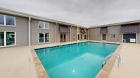 Located at 1000 N Donahue Dr in Auburn, Alabama, this outdoor amenity area centers around a generously sized swimming pool with crystal-clear blue water and convenient entry handrails on multiple sides. The pool is flanked by a wide, light-colored concrete sun deck that provides ample space for relaxation and recreation. Rows of sling-style lounge chairs are arranged throughout the deck, offering abundant seating for residents or guests. A covered pergola structure at one end of the deck provides a shaded gathering spot, complemented by additional patio furniture for outdoor dining and socializing. The pool area is fully enclosed by a black metal security fence with a gated entry, ensuring a safe and controlled environment. An adjacent two-story brick and stucco amenity building borders one side of the pool, featuring metal awnings over its doors and windows and direct access points to the pool deck. The building's exterior is finished in a neutral tone that complements the surrounding hardscape. Mature trees and natural landscaping are visible beyond the fence line, providing a green backdrop. The entire space is thoughtfully designed to serve as a comprehensive outdoor leisure destination, combining swimming, sunbathing, and social amenities in a well-organized, attractive setting.