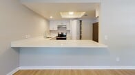 This single-floor residence offers a clean, functional layout with a neutral color palette and a variety of thoughtful finishes throughout. The open-concept living and dining area is finished with light wood-look flooring and features multiple windows fitted with horizontal blinds, allowing natural light to fill the space. A textured ceiling adds subtle character to the room, while a pass-through breakfast bar connects the living area seamlessly to the kitchen.

The kitchen is well-appointed with white cabinetry, a stainless steel range, a built-in sink, and light-toned countertops that extend to form the bar-height peninsula. A tray ceiling detail above the kitchen adds architectural interest, and ample cabinet storage is complemented by a pantry closet.

The two bedrooms are carpeted in a warm beige tone and share a similarly neutral aesthetic, each featuring a window with blinds that provides natural light and ventilation. The rooms offer generous wall space suitable for a variety of furniture arrangements.

The bathroom is finished with light wood-look flooring and includes a tiled tub-and-shower combination with a curved curtain rod, a toilet, and a white vanity with under-sink storage. Clean lines and a bright, simple design give the bathroom a fresh, functional character. Overall, the space presents a comfortable and practical living environment.