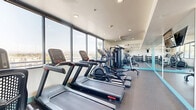 Located in Escondido, California, this fitness center offers a thoughtfully designed exercise space built to support a well-rounded workout routine. The room is laid out to maximize functionality, with dedicated zones for both cardio and strength training. Along one side, a row of treadmills and elliptical machines lines the large picture windows, which flood the space with natural light and provide scenic views of the surrounding Southern California landscape. A full-wall mirror runs along the opposite side, mounted with a flat-screen television and serving to visually expand the room while allowing users to monitor their form during workouts. The strength training area features a multi-station cable machine alongside a tiered dumbbell rack stocked with a wide range of free weights, complemented by exercise balls for functional training. Recessed ceiling lighting provides even illumination throughout, while a ceiling fan supports air circulation during intense sessions. The floor is finished with durable rubber gym flooring in a neutral gray tone, accented by teal trim along the base of the walls, lending the space a clean, modern aesthetic. The overall design balances practicality with a bright, inviting ambiance well-suited for daily fitness use.