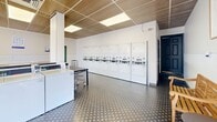 Located at 8650 Kingsbridge Dr in University City, Missouri, this community laundry facility offers a clean, functional, and thoughtfully designed space for residents. The single-floor layout provides a comfortable amount of room to move freely between machines and folding areas.

The facility is equipped with a substantial row of front-load washers along one wall and a full bank of stacked and standalone dryers along the opposite wall, providing ample capacity for simultaneous use. A centrally positioned folding table offers a dedicated surface for sorting and folding laundry with ease.

The interior design balances utility with comfort. Decorative patterned tile flooring adds a stylish touch throughout, while a warm wood-slat ceiling paired with recessed panel lighting creates an inviting ambiance uncommon in laundry facilities. White-painted walls keep the space feeling bright and clean.

Natural light is introduced through a large glass block window, which diffuses soft, even light across the room without direct glare. A glass-panel entry door further enhances the brightness of the space. Wooden benches and chairs are positioned near the entrance and along the walls, providing comfortable seating for patrons waiting on their laundry. Informational signage is posted throughout, and a fire extinguisher and exit signage ensure safety compliance. The overall atmosphere is tidy, well-maintained, and welcoming.