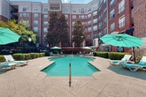 This outdoor amenity space is centered around a generously sized freeform swimming pool with clear turquoise water, situated within a semi-enclosed courtyard formed by a multi-story red brick building. The building's curved and angular facade creates a sense of enclosure, providing a sheltered atmosphere while still allowing ample natural light and open sky above.

The pool deck is finished in a warm stamped concrete surface, offering a spacious surround on all sides. Multiple lounge chairs and chaise lounges are positioned throughout the deck, accompanied by teal-canopied umbrellas that provide shade at various points around the pool. The layout accommodates both sun and shade preferences throughout the day.

The courtyard is enhanced by mature trees, including prominent magnolias, which add natural greenery and additional shade. Neatly trimmed hedgerows and ornamental plantings border the perimeter, contributing to the well-maintained aesthetic. A wrought iron fence delineates the boundary of the pool area, adding a classic architectural detail consistent with the building's traditional brick exterior. Wall-mounted and freestanding light fixtures are present throughout, ensuring the space is functional beyond daylight hours. Overall, the courtyard presents a refined, resort-style outdoor environment.