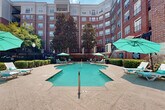 This outdoor amenity space is centered around a generously sized freeform swimming pool with clear turquoise water, situated within a semi-enclosed courtyard formed by a multi-story red brick building. The building's curved and angular facade creates a sense of enclosure, providing a sheltered atmosphere while still allowing ample natural light and open sky above.

The pool deck is finished in a warm stamped concrete surface, offering a spacious surround on all sides. Multiple lounge chairs and chaise lounges are positioned throughout the deck, accompanied by teal-canopied umbrellas that provide shade at various points around the pool. The layout accommodates both sun and shade preferences throughout the day.

The courtyard is enhanced by mature trees, including prominent magnolias, which add natural greenery and additional shade. Neatly trimmed hedgerows and ornamental plantings border the perimeter, contributing to the well-maintained aesthetic. A wrought iron fence delineates the boundary of the pool area, adding a classic architectural detail consistent with the building's traditional brick exterior. Wall-mounted and freestanding light fixtures are present throughout, ensuring the space is functional beyond daylight hours. Overall, the courtyard presents a refined, resort-style outdoor environment.