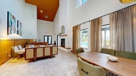 Located at 2201 San Jose Dr in Antioch, California, this thoughtfully designed single-floor space offers an open, airy layout that makes a strong architectural impression. The most striking feature is the dramatically vaulted ceiling clad in warm wood paneling, which rises to an impressive height and floods the interior with natural light through strategically placed clerestory windows and large glass doors.

The combined living and dining area forms the heart of the space. The living zone features a clean-lined fireplace framed by a wood mantel and an arched mirror above, flanked by warm wall sconces. Comfortable seating areas are accented by a wood-paneled wainscoting wall, a checkered area rug, and vibrant contemporary artwork, creating a welcoming, eclectic atmosphere. The dining area accommodates a solid wood oval table surrounded by upholstered green chairs, positioned beside a bold black-framed glass partition wall that connects to an adjacent room.

The kitchen is distinctly styled with terracotta-red shaker cabinets, stone countertops, open wood shelving, and a large window that brings in natural light and outdoor views. Terrazzo-style tile flooring flows consistently throughout the space, tying the rooms together cohesively. Large glass doors open to views of an outdoor pool area, further enhancing the bright, connected feel of the interior.
