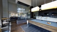The Clubroom at 100 Danforth Ave in Dobbs Ferry, NY is a sophisticated, single-floor amenity space designed for social gatherings and leisure. The interior immediately impresses with soaring double-height ceilings and an open, airy layout that creates a grand sense of space. A centerpiece billiards table with a richly grained wood frame anchors the main recreation area, set atop a striped area rug over sleek hardwood-style flooring. Dramatic pendant lighting with oversized rectangular shades illuminates the space with a warm, modern glow. A striking floor-to-ceiling glass enclosure houses a contemporary fireplace feature, serving as both a visual focal point and a divider between zones. Adjacent to the recreation area, a lounge and dining section offers stylish seating arrangements ideal for casual gatherings. A white kitchenette with bar-height seating provides a convenient serving and social hub. Large windows throughout the space invite abundant natural light and frame views of the surrounding greenery. Dark cabinetry and trim details contrast elegantly with lighter surfaces, reinforcing the space's upscale, contemporary aesthetic. The overall ambiance is refined yet inviting, making it a premier gathering destination.