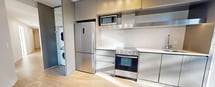 This thoughtfully designed two-bedroom, two-bathroom residence offers a clean, contemporary aesthetic throughout its efficiently organized single-floor layout. Light-toned wood-style flooring runs continuously through the living areas and bedrooms, creating a cohesive, modern feel.

The kitchen is a standout feature, outfitted with sleek stainless steel appliances including a full-size refrigerator, oven with range, built-in microwave, and a stainless steel range hood. Flat-panel cabinetry in a muted metallic finish extends to the ceiling for generous storage, complemented by white countertops and a large floor-to-ceiling window that floods the space with natural light and provides views of the surrounding urban landscape.

The bedrooms are bright and airy, with crisp white walls and large windows that allow sunlight to cast across the light flooring. Both bathrooms are finished with neutral tile flooring, white soaker tub-shower combinations, modern floating vanities with white countertops, and large mirrors with chrome fixtures throughout.

The unit also includes a dedicated laundry area, a closet, and multiple hallways for practical circulation. A spacious outdoor balcony with glass railings offers open views of the neighboring cityscape under wide, clear skies — an ideal spot for enjoying the outdoors. The overall ambiance is clean, minimalist, and move-in ready.