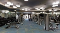 This modern fitness center is designed to accommodate a full range of workout routines within a single, open-plan floor. The space is outfitted with an extensive selection of strength training machines, free weights, and cable equipment, providing users with everything needed for a complete workout. Large floor-to-ceiling mirrors line the walls, enhancing the sense of space and allowing users to monitor their form during exercise. The flooring features a durable, patterned carpet in teal and gold tones, adding a polished aesthetic while providing comfort underfoot. Overhead, a series of large, round pendant light fixtures are suspended from a dark tray ceiling, casting even, bright illumination throughout the space. Natural light filters in through glass doors at the far end of the room, connecting the interior to the outside and contributing to an open, airy ambiance. The layout is thoughtfully organized to allow ample circulation between equipment stations. The overall design strikes a balance between functionality and contemporary style, making it a well-appointed facility suitable for a wide range of fitness needs.