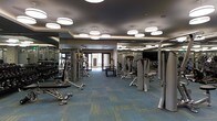 This modern fitness center is designed to accommodate a full range of workout routines within a single, open-plan floor. The space is outfitted with an extensive selection of strength training machines, free weights, and cable equipment, providing users with everything needed for a complete workout. Large floor-to-ceiling mirrors line the walls, enhancing the sense of space and allowing users to monitor their form during exercise. The flooring features a durable, patterned carpet in teal and gold tones, adding a polished aesthetic while providing comfort underfoot. Overhead, a series of large, round pendant light fixtures are suspended from a dark tray ceiling, casting even, bright illumination throughout the space. Natural light filters in through glass doors at the far end of the room, connecting the interior to the outside and contributing to an open, airy ambiance. The layout is thoughtfully organized to allow ample circulation between equipment stations. The overall design strikes a balance between functionality and contemporary style, making it a well-appointed facility suitable for a wide range of fitness needs.