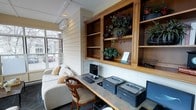 This single-floor space functions as a compact yet well-equipped business center with a warm, residential aesthetic. The room features white shiplap-style wall paneling that lends a clean, cottage-inspired character throughout. A built-in wall-mounted shelving unit crafted from warm-toned wood dominates one wall, offering multiple open compartments adorned with lush decorative greenery and potted plants, adding a natural, inviting touch to the functional setting.

A continuous countertop workstation runs along the wall beneath the shelving, providing ample desk space accommodating a desktop computer with keyboard and mouse, as well as multiple laser printers, making the area well-suited for business and administrative tasks. A wooden chair with an upholstered seat is positioned at the workstation for comfortable use.

To one side, a cream-colored upholstered sofa with accent pillows creates a relaxed seating area, softening the workspace atmosphere. Natural light filters in through a large window overlooking an outdoor street view, brightening the room. A wall-mounted mini-split air conditioning unit ensures year-round climate comfort. Overall, the space balances practicality with a cozy, approachable ambiance.